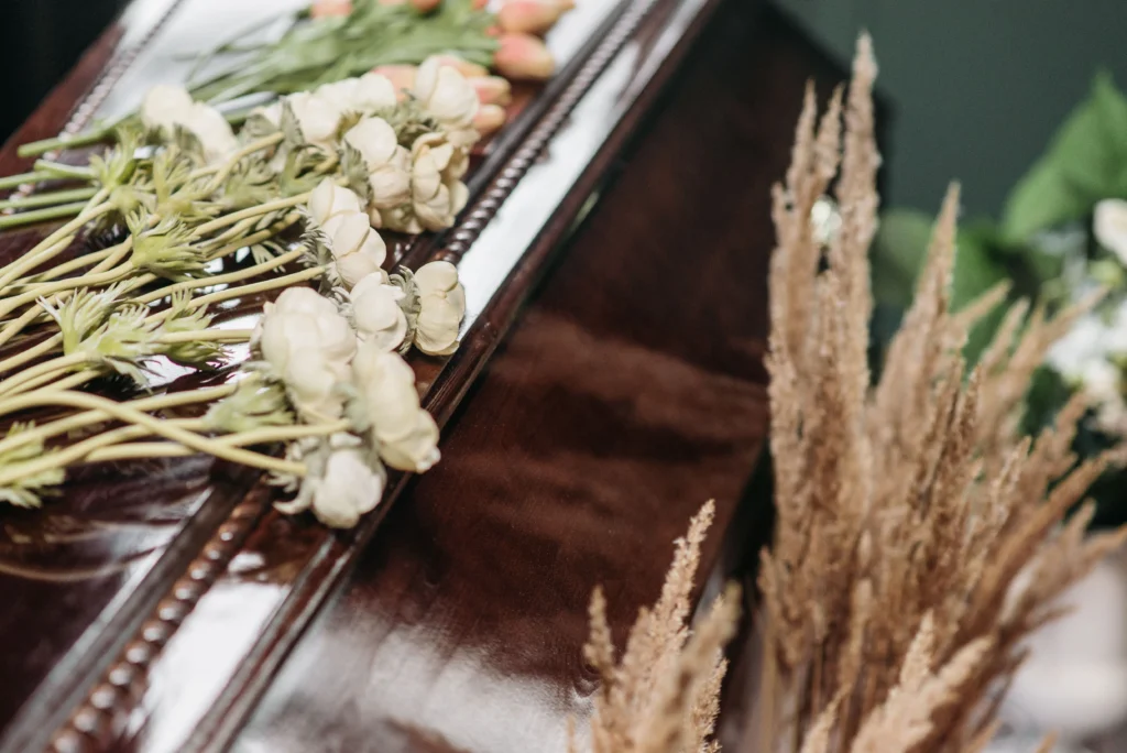Coffin-pexels-pavel-danilyuk-7317674 - Insider Accident Lawyers A solemn funeral arrangement featuring a wooden casket adorned with white ranunculus and dried grasses.