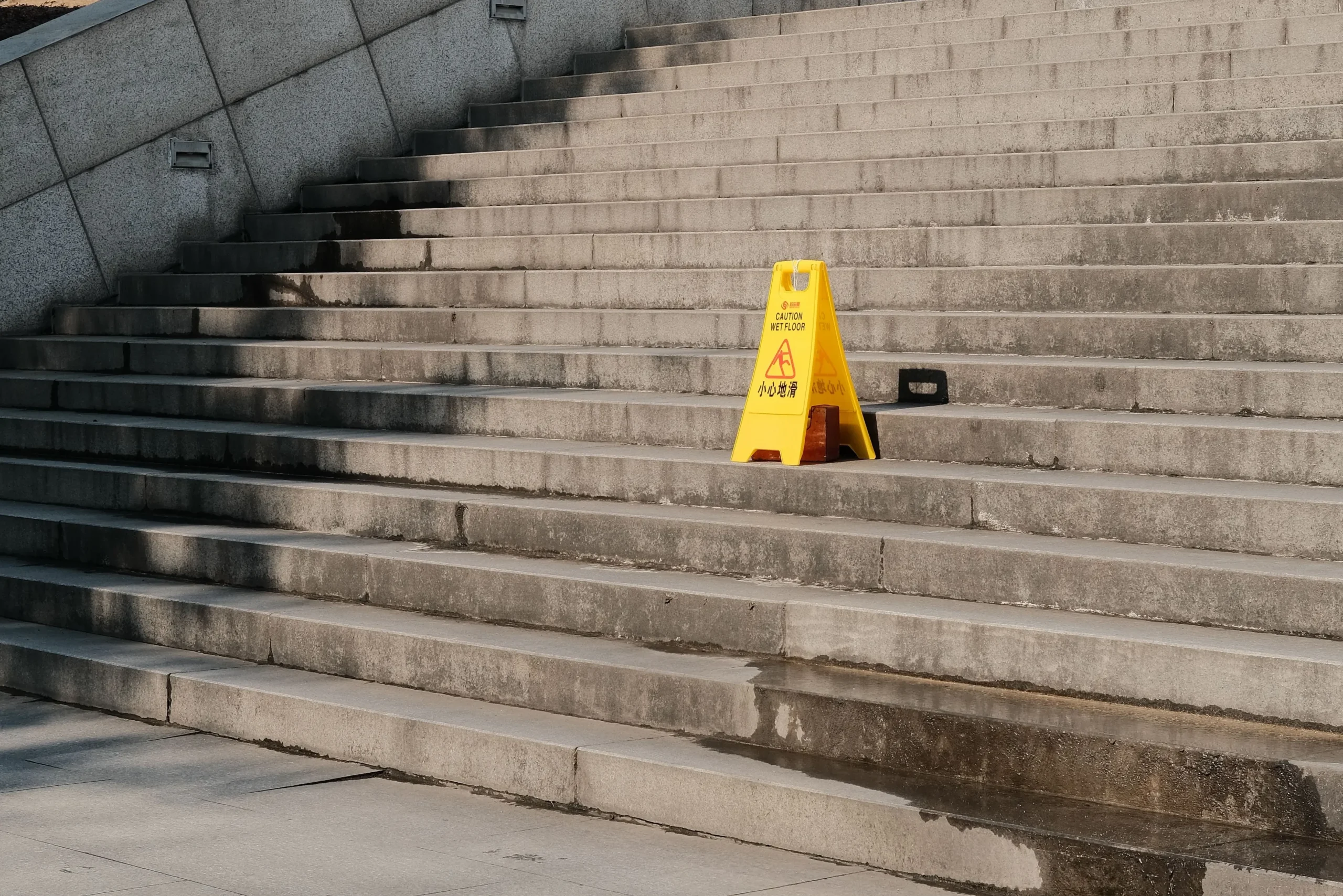 pexels-tizzy-30881646 - Insider Accident Lawyers Concrete outdoor stairs with a bright yellow warning sign reading'Caution Wet Floor' on the lower steps; the ground is wet with reflections from puddles.